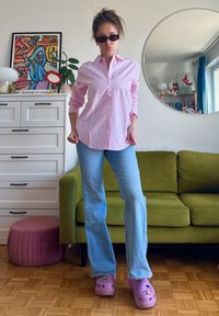 Camisa rosa às riscas, calças de ganga azul clarinho em forma de sino e tamancos roxos com buracos decorativos. Sofá verde e espelho redondo ao fundo.