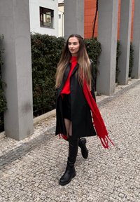 Mustast nahast mantel, punane kampsun ja sall, kõrge vöökohaga lühikesed püksid ning põlveni ulatuvad mustad saabad tekstuurse tänavapõranda peal. Taustal põõsad.