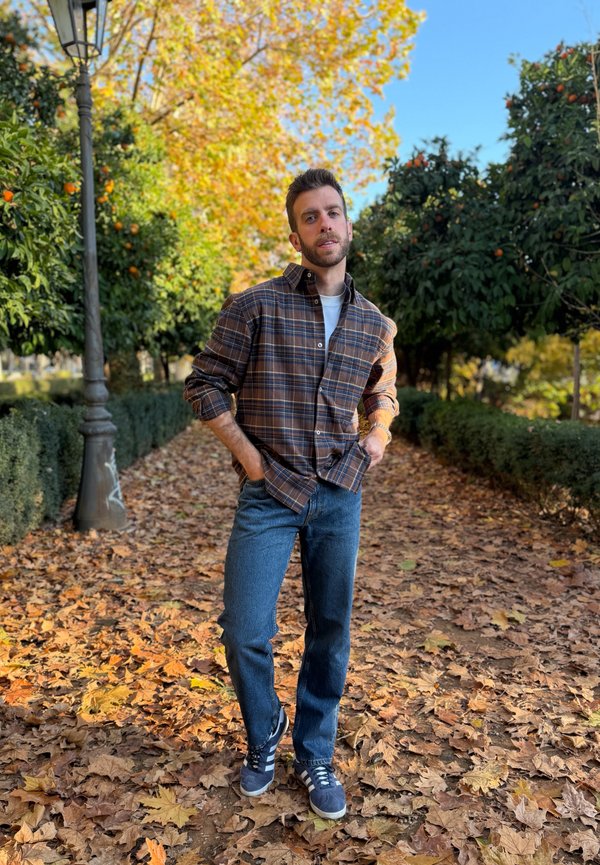 Homme en chemise à carreaux et jeans debout sur un chemin couvert de feuilles entre des orangers sous un ciel d'automne.