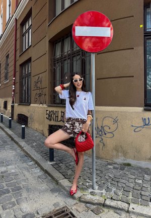 Camiseta blanca con un pequeño gráfico, pantalones cortos marrón con estampado, bolso texturizado rojo, zapatos planos rojos y gafas de sol, posando cerca de una señal de entrada prohibida.
