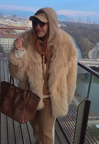 Femme portant un manteau en fourrure beige, un sweat à capuche, une casquette, des lunettes de soleil et un pantalon en tricot, tenant un grand sac marron sur un balcon avec vue sur la ville et la rivière.