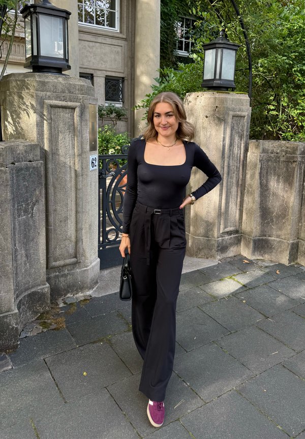 Zwarte lange mouw top met vierkante halslijn, hoog uitgesneden zwarte broek en paarse sneakers; geaccessoireerd met een kleine zwarte handtas.