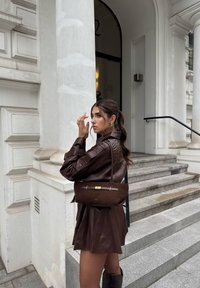 Bruine suède handtas met een zachte textuur, voorzien van een gouden accent sluiting en een zachte rechthoekige vorm. Draag hem bij een bruine leren outfit.