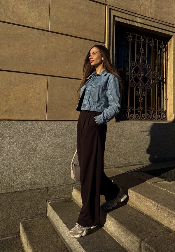 Donna con i capelli lunghi che indossa una giacca di jeans, pantaloni neri e scarpe da ginnastica, in piedi su scale esterne accanto a un muro di pietra e a una finestra decorativa in ferro.