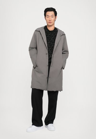 Abrigo largo gris con capucha, hecho de material suave, con botones en la parte frontal y bolsillos laterales. Lleva puesto un suéter negro texturizado y pantalones negros.