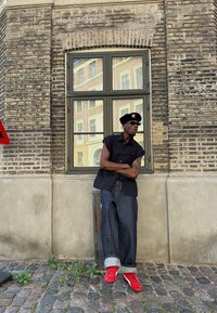 Μαύρο αμάνικο μπλουζάκι, υπερμεγέθεις τζιν με γυρισμένα μπατζάκια και κόκκινα αθλητικά παπούτσια. Σταθείτε μπροστά σε έναν υφασμάτινο τοίχο από τούβλα κοντά σε ένα παράθυρο.