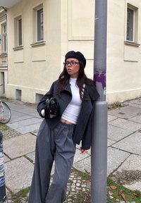 Šedý vlněný kabát přes bílý top, ve spojení se širokými šedými kalhotami. Doplňky zahrnují černou kabelku a černý baret, na neutrálním pozadí.