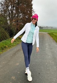 Bijela preklopna jakna, svijetloplava majica, mornarsko plave tajice, ružičasta kapa, bijele tenisice. Hoda po stazi s zelenom travom i drvećem u pozadini.
