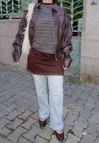 Žena v hnědé kožené bundě, pruhovaném tričku a hnědé sukni přes světle modré džíny, nesoucí bílou chlupatou kabelku, stojí na dlážděném chodníku.