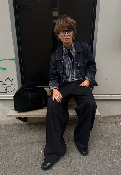 Chaqueta de mezclilla sobre una camisa de rayas, pantalones anchos negros y zapatos negros. Bolsa negra al lado de la persona sentada en un banco de madera.