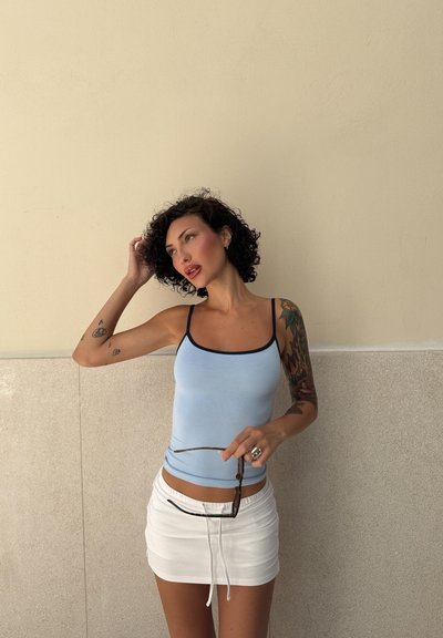Top sin mangas ajustado de color azul claro con ribete azul marino, combinado con una falda corta blanca. Sostiene gafas de sol, mostrando coloridos tatuajes en el brazo.
