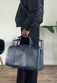 Tmavomodrá kožená kabelka s hladkou textúrou, zaobleným tvarom, dvoma hornými rúčkami a striebornými kovaniami. Na prednej strane je logo.