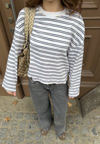 Chemise blanche à manches longues avec rayures bleu marine, coupe décontractée ; associée à un jean gris ample et des chaussures marron ; portant un sac à main beige tissé.