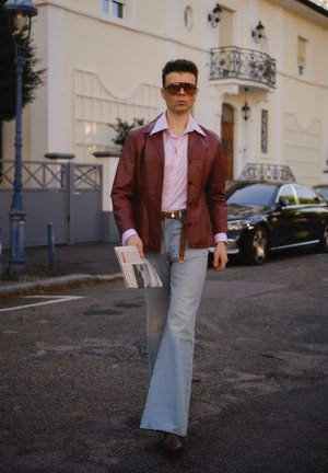 Chaqueta de cuero roja, camisa rosa clara, jeans claros de campana, zapatos marrones. Sosteniendo un periódico, caminando por una calle con un coche al fondo.