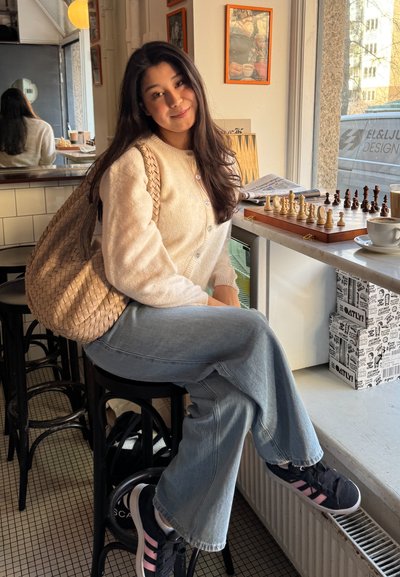 Cardigan crema, jeans acampanados azul claro y zapatillas negras con rayas rosas. Una bolsa de mimbre reposa contra una silla en un ambiente de café.