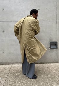 Άνδρας με ταμπά καμπαρντίνα, γκρι παντελόνι και μαύρα παπούτσια στέκεται με την πλάτη γυρισμένη μπροστά σε έναν τσιμεντένιο τοίχο με ένα στόμιο εξαερισμού.