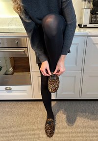 Leopardenmuster-Slipper mit schwarzen Schnürsenkeln, getragen mit schwarzen Leggings, während die Person den Schuh in einer Küchenszene anpasst.