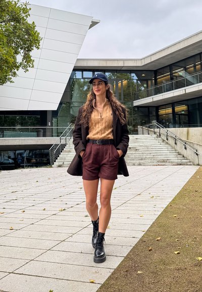 Abrigo marrón sobre un suéter de punto beige, pantalones cortos burdeos y botas negras con cordones. De pie sobre un camino de piedra con arquitectura moderna detrás.