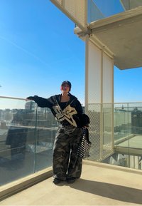 Hoodie preto oversized com gráfico branco, calças baggy camufladas, ténis pretos e um boné preto. Segurando uma mala com tachas. Céu azul brilhante.