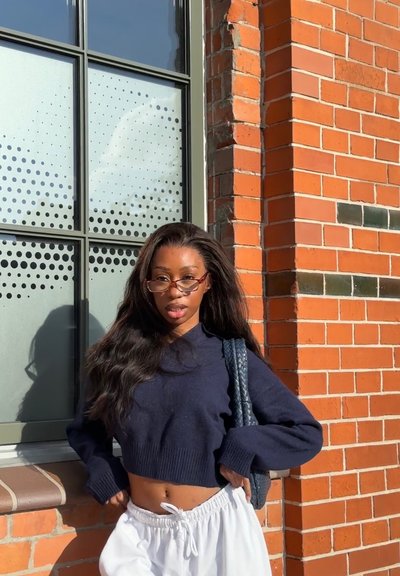 Suéter corto navy con dobladillo acanalado, pantalones blancos con cordón y bolso trenzado, contra un fondo de ladrillo texturizado y una ventana.