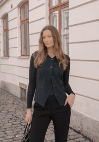 Colete preto com botão na frente, combinado com uma blusa de mangas longas. O modelo está de pé contra uma parede clara com janelas, segurando uma mala.