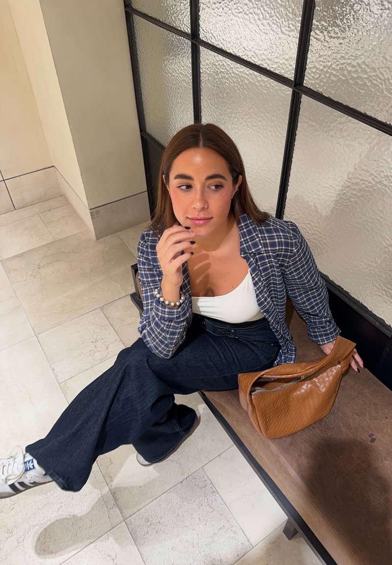 Une personne est assise sur un banc en bois, portant une chemise en flanelle bleue à carreaux, un haut blanc, un jean foncé et des baskets blanches, avec un sac à main en cuir brun à côté d'elle.