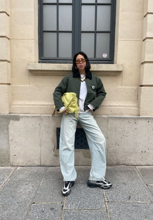 Persona con chaqueta verde, camisa blanca, vaqueros anchos de pierna azul claro, zapatillas blancas y negras, sosteniendo una bolsa amarilla, de pie frente a una pared de piedra beige.