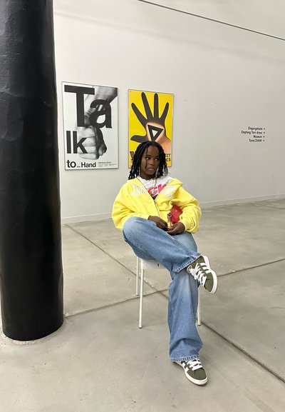 Joven sentado en una silla blanca, vistiendo una sudadera amarilla con detalles gráficos, jeans de mezclilla azul claro y zapatillas verdes. Pilar negro cerca.