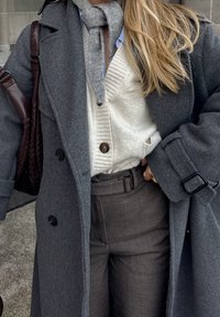 Femme portant plusieurs couches de vêtements d'hiver, y compris un manteau en laine gris, un gilet crème, une chemise bleue, une écharpe grise, un pantalon marron, et portant un sac tissé marron.