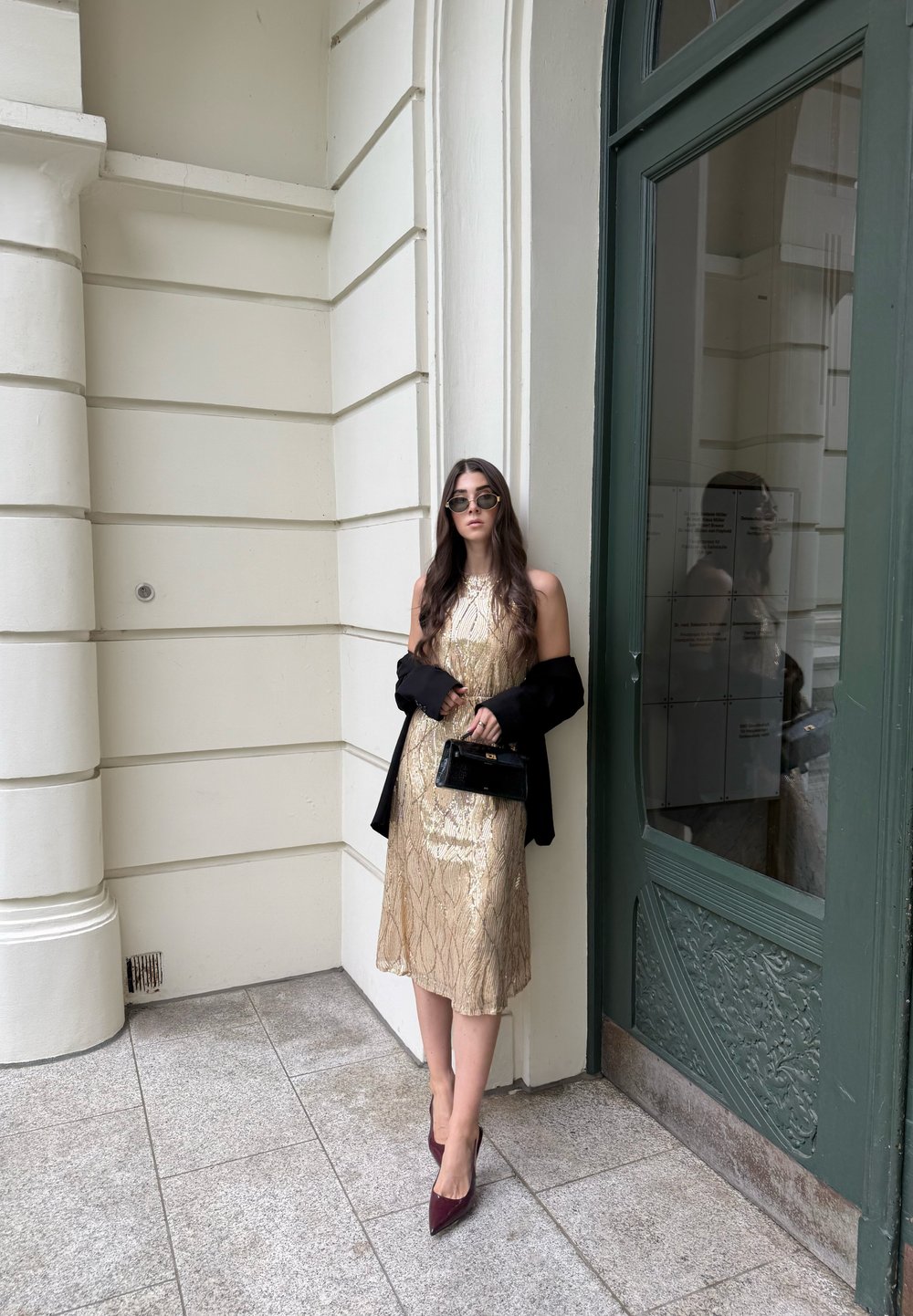 Mujer con cabello largo vistiendo un vestido dorado, chaqueta negra sobre los brazos, gafas de sol, sosteniendo un pequeño bolso negro, de pie junto a una puerta verde.