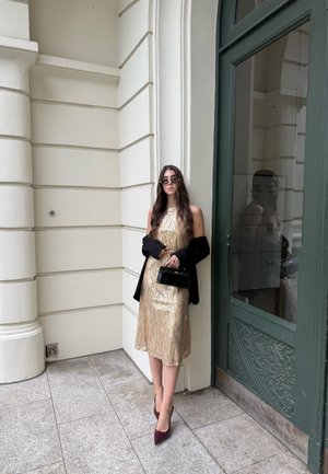 Mujer con cabello largo vistiendo un vestido dorado, chaqueta negra sobre los brazos, gafas de sol, sosteniendo un pequeño bolso negro, de pie junto a una puerta verde.