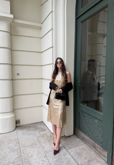 Mujer con cabello largo vistiendo un vestido dorado, chaqueta negra sobre los brazos, gafas de sol, sosteniendo un pequeño bolso negro, de pie junto a una puerta verde.
