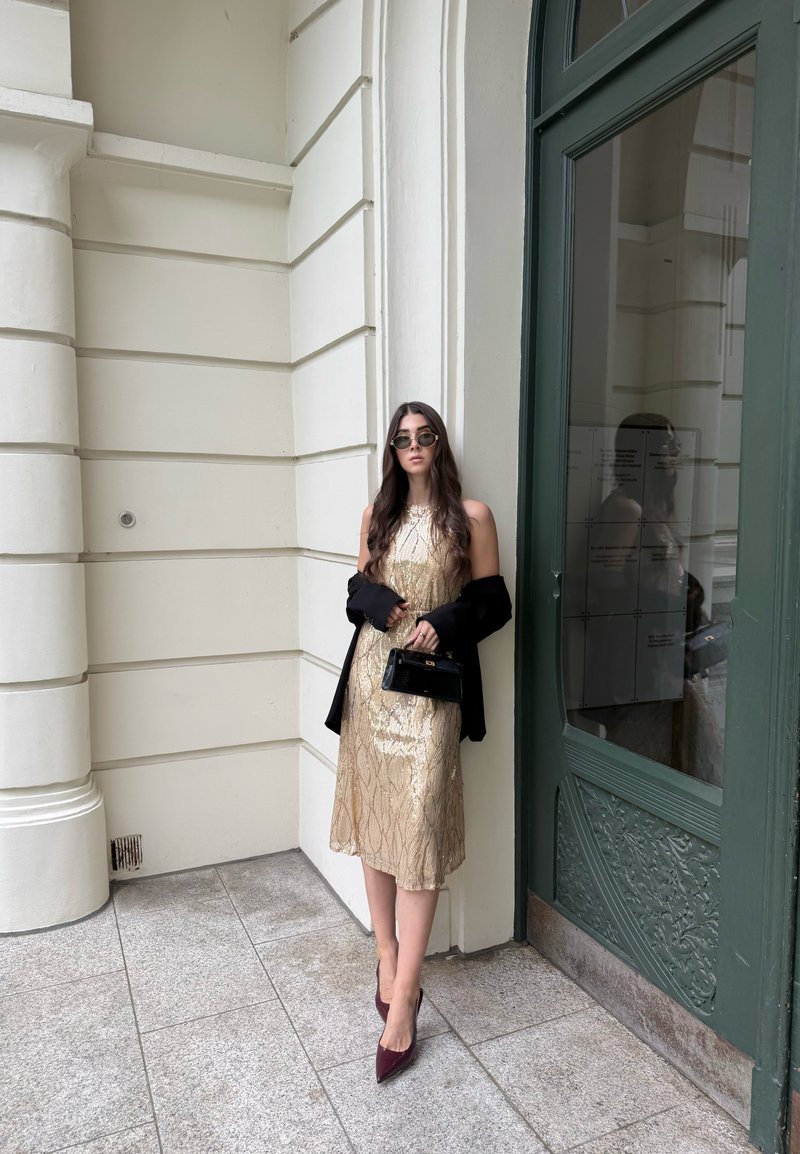 Frau mit langen Haaren, trägt ein goldenes Kleid, schwarze Jacke über den Armen, Sonnenbrille, hält eine kleine schwarze Handtasche, steht vor einer grünen Tür.