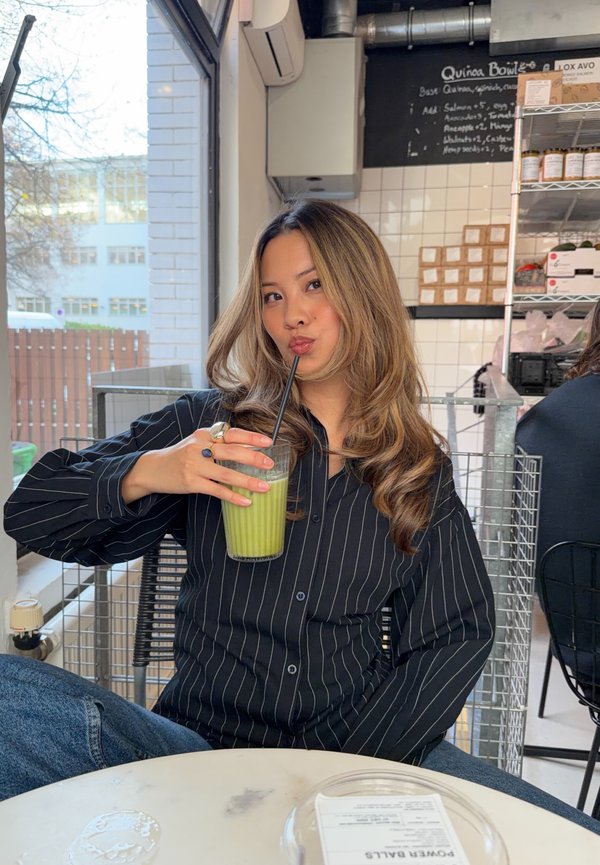 Chemise noire à rayures fines à boutons, verre de boisson verte avec une paille, table ronde, décor industriel de café, lumière naturelle à travers les fenêtres.