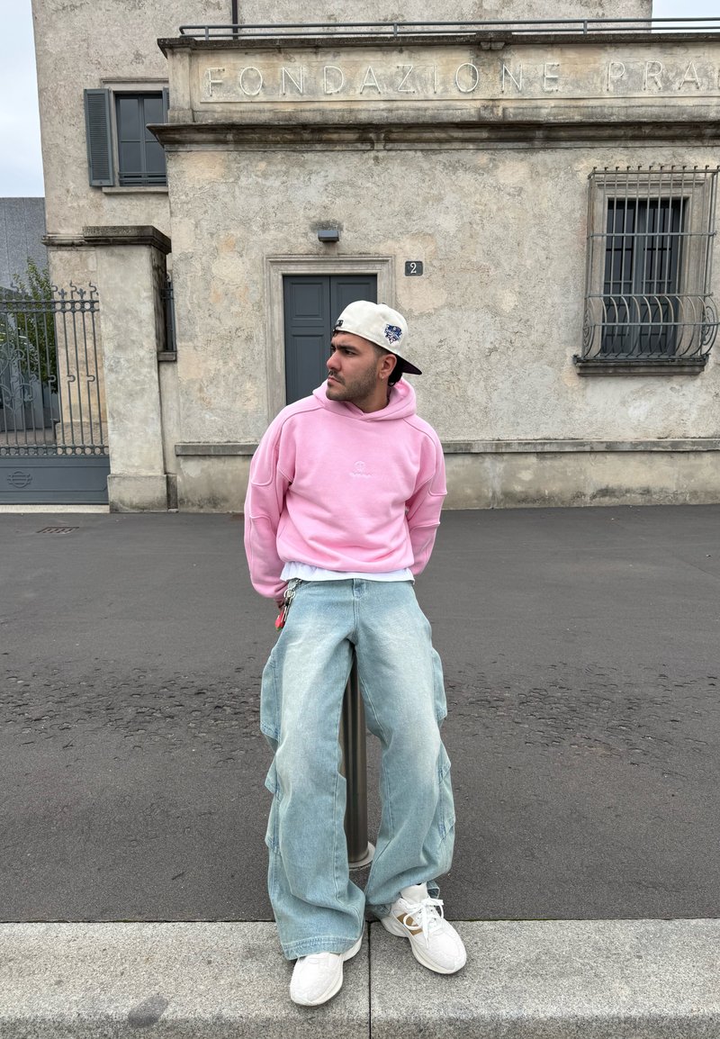 Ροζ oversize φούτερ, φαρδιά ανοιχτό μπλε τζιν με πλαινές τσέπες, λευκά αθλητικά παπούτσια και λευκό καπέλο με μπλέ λεπτομέρειες. Αστική τοποθεσία.