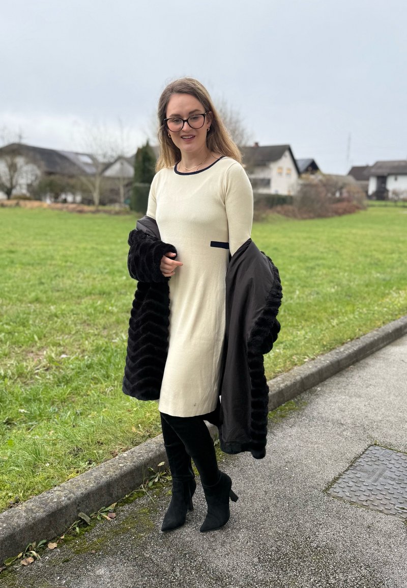 Cream-colored dress with navy accents, paired with a black textured coat. The wearer has black knee-high boots and stands on a grassy path.