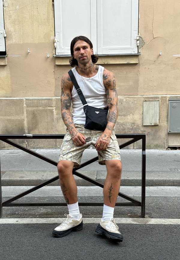 Mand med tatoveringer iført hvid tanktop, mønstrede shorts, hvide sokker og sneakers sidder på en metalbænk på en byfortov.