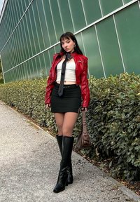 Jaqueta de couro vermelha, top curto branco, saia mini preta, botas altas pretas até ao joelho, gravata slim, a segurar uma mala castanha, de pé ao ar livre.