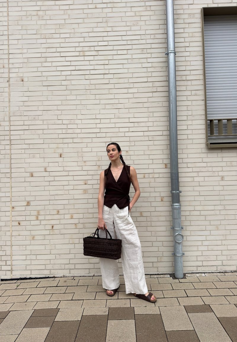 Donna con capelli intrecciati che indossa un top scuro senza maniche e pantaloni bianchi larghi, in piedi accanto a un muro di mattoni beige tenendo una borsa scura intrecciata.