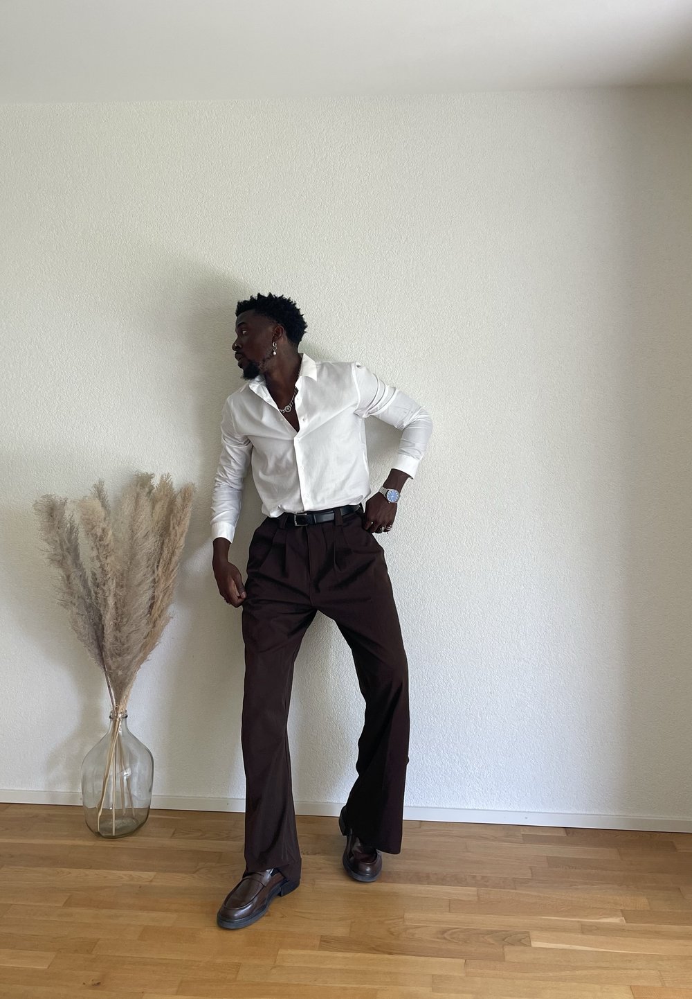 Weißes Langarmhemd mit lockerer Passform, braune weite Hosen und dunkle Schuhe; Pampasgras in einer klaren Vase in der Nähe.