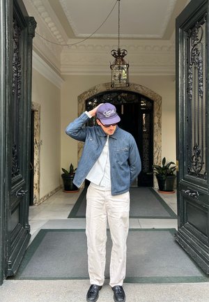 Hombre con gafas de sol y gorra ajusta su sombrero mientras está de pie en una entrada ornamentada con puertas dobles negras abiertas y plantas enmacetadas dentro.