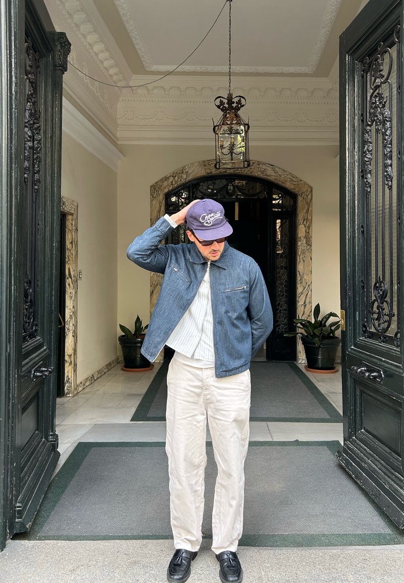Homme en lunettes de soleil et casquette ajuste son chapeau devant une entrée ornée avec des portes doubles noires ouvertes et des plantes en pot à l'intérieur.