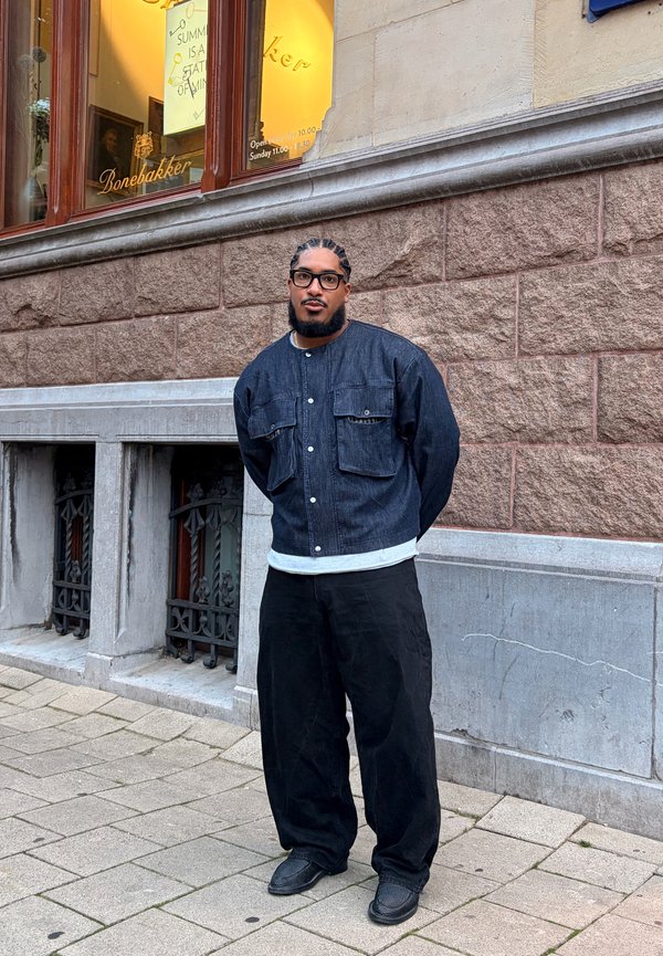 Homme avec des cheveux tressés et des lunettes portant une veste en denim sombre et un pantalon noir se tient sur un trottoir en pierre près d'un mur en briques.