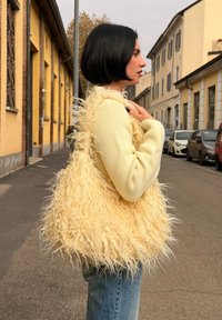Borsa tote gialla e soffice realizzata in tessuto texturizzato, caratterizzata da lunghe fibre morbide. La borsa è indossata a spalla contro uno sfondo urbano.