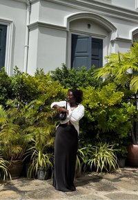 Kvinne i en hvit bluse og lang svart skjørt som justerer svart veske, stående på steinbelagt fortau foran grønne planter og grå bygningdører.