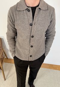 Άτομο που φοράει ελαφρώς καφέ ριμπ πλεκτή ζακέτα με κουμπιά πάνω από μαύρο πουκάμισο, με μαύρο παντελόνι και χέρια στις τσέπες, στέκεται μέσα σε εσωτερικό χώρο πάνω σε χαλί.