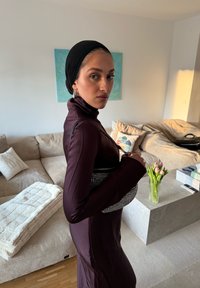 Femme en robe violet foncé et foulard noir tenant un sac à main clouté, debout dans un salon avec canapé beige et tulipes sur une table en marbre.