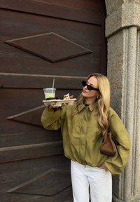 Masivna jakna maslinastozelene boje, bijele hlače, drži pladanj s pićem i pecivom. U pozadini je tamna drvena vrata s uzorkom u obliku dijamanta.