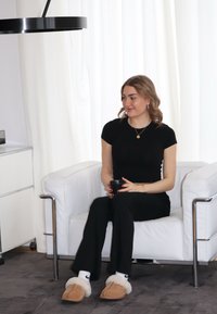 Λευκή πολυθρόνα με μεταλλικά πόδια, ένα άτομο σε μαύρη ενδυμασία και παντόφλες κρατά ένα μαύρο φλιτζάνι, καθισμένο σε ένα γκρίζο χαλί με ανοιχτό φόντο.