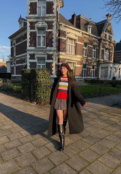Mujer con abrigo negro, suéter de rayas, falda gris y botas caminando por la acera frente a un gran edificio de ladrillo y piedra.